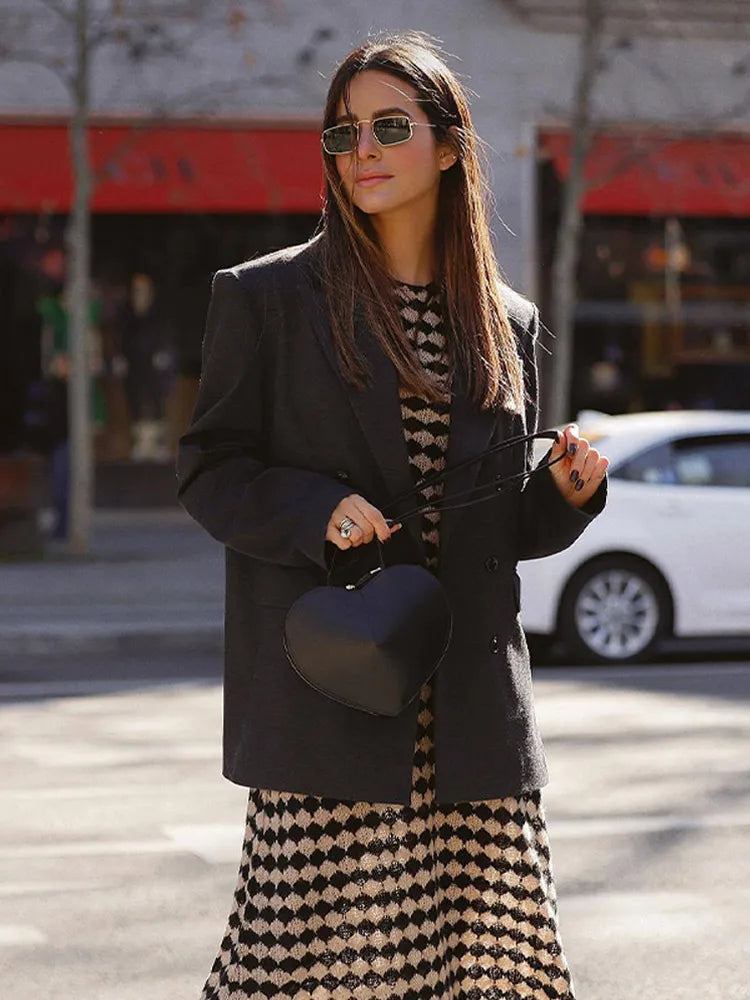 Woman wearing a black blazer over a knit dress with a black and beige geometric diamond pattern, carrying a heart-shaped black handbag.