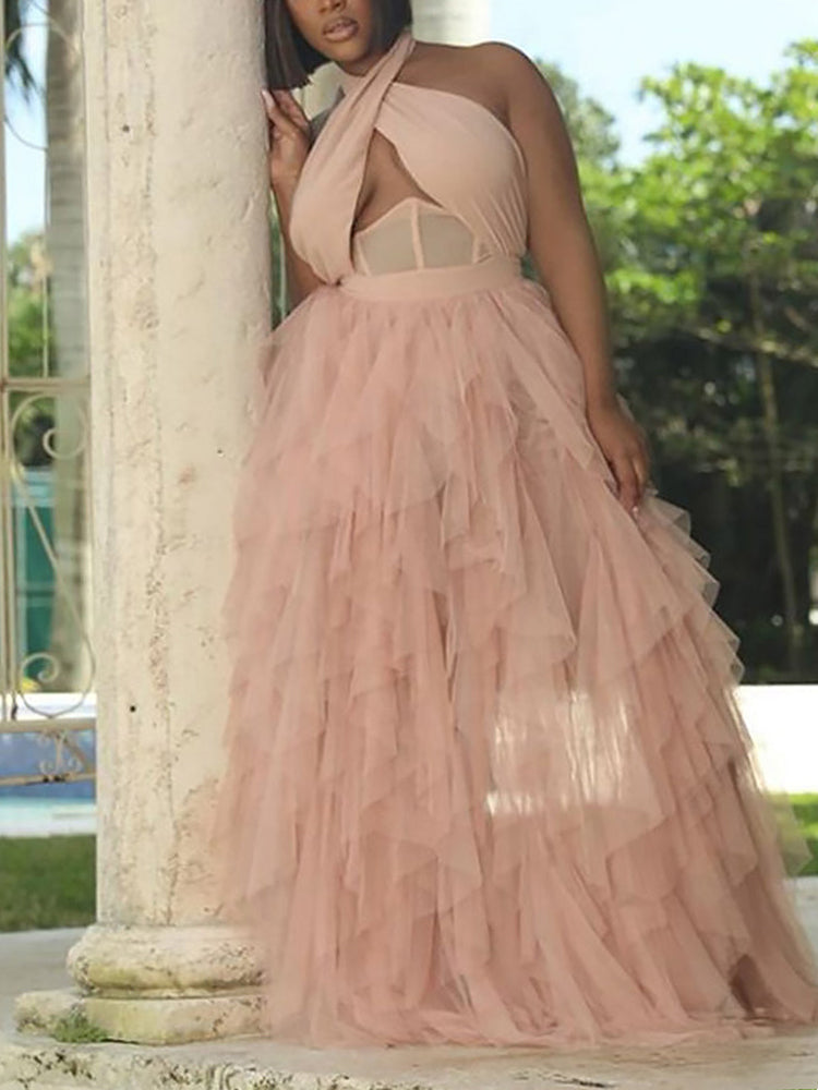 Woman leaning against a pillar in a blush pink halter neck maxi dress with a tiered tulle skirt, holding a basket of flowers.
