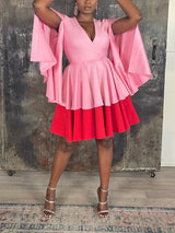 Woman in a pink and red color-block dress with cape sleeves and a flared skirt, standing in front of a rustic metal door.