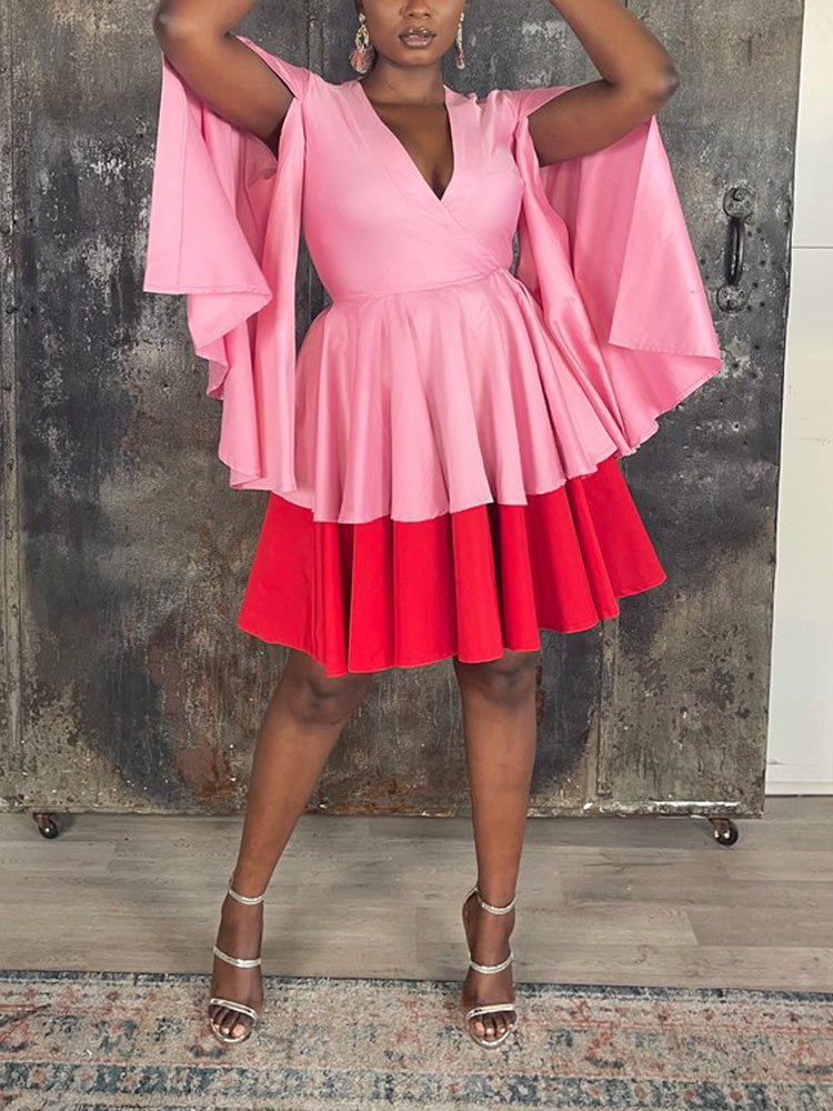 Woman in a pink and red color-block dress with cape sleeves and a flared skirt, standing in front of a rustic metal door.