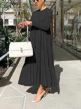 Woman holding a white quilted handbag while wearing a long, black tiered maxi dress with long sleeves.