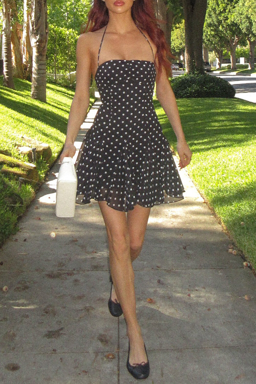 Woman in a black and white polka dot halter dress and black ballet flats, holding a white handbag.