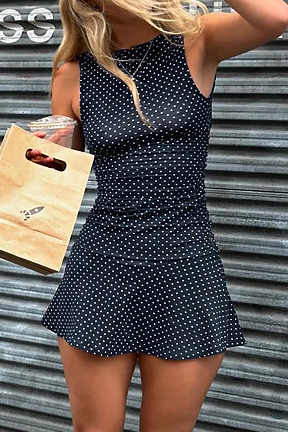 Woman holding a drink and shopping bag, wearing a navy polka dot mini dress.