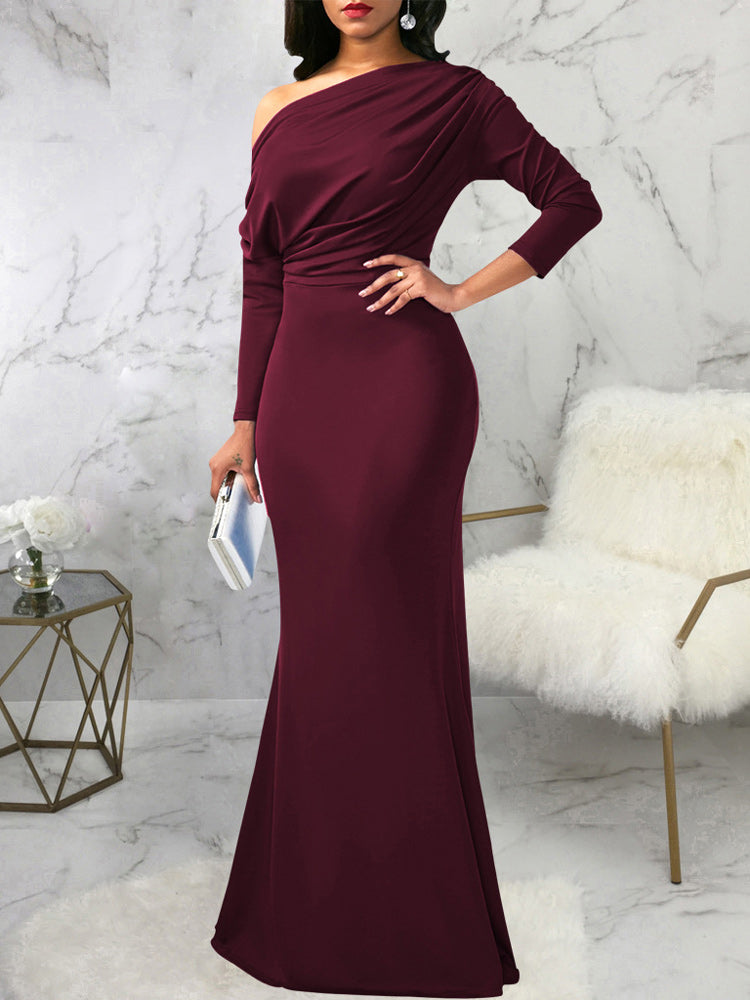 Woman wearing a long burgundy asymmetrical dress with long sleeves and a mermaid hemline, holding a clutch.
