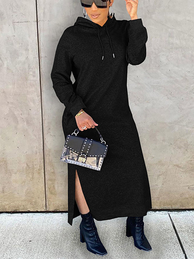 Woman holding a black handbag, wearing a long black hooded dress with side slits, and black boots.