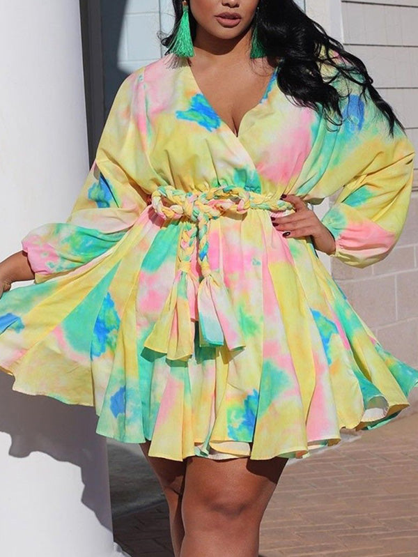 Close-up of a plus-size woman wearing a yellow tie-dye mini dress with a V-neck, belted waist, and full sleeves, with green tassel earrings.