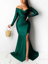 Woman wearing a long, emerald green, off-the-shoulder satin gown with a high slit and mermaid hemline, holding a gold clutch.