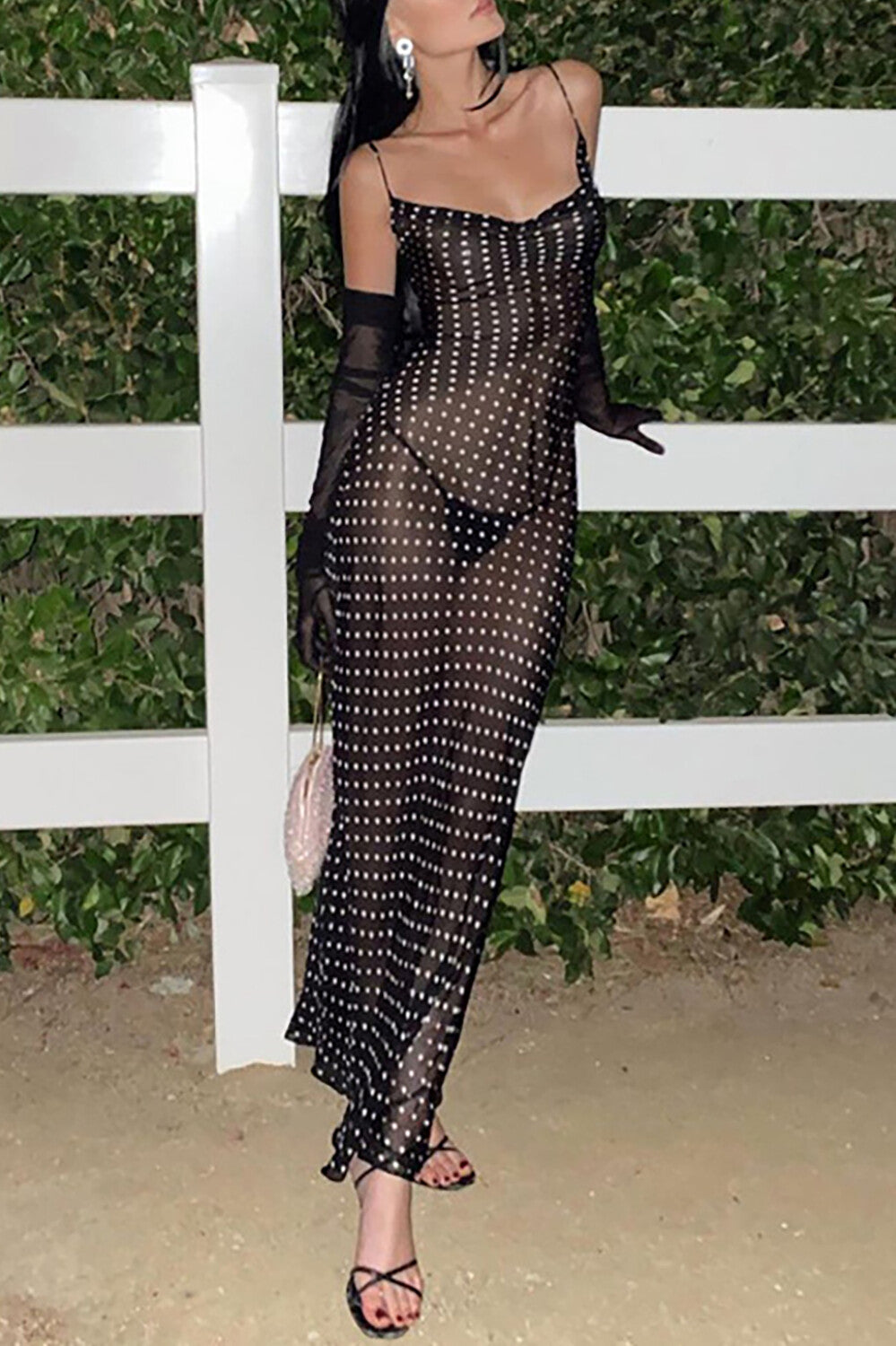 Woman wearing a black polka dot sheer maxi dress with spaghetti straps and a cowl neck, with black sheer gloves and a small pink handbag.