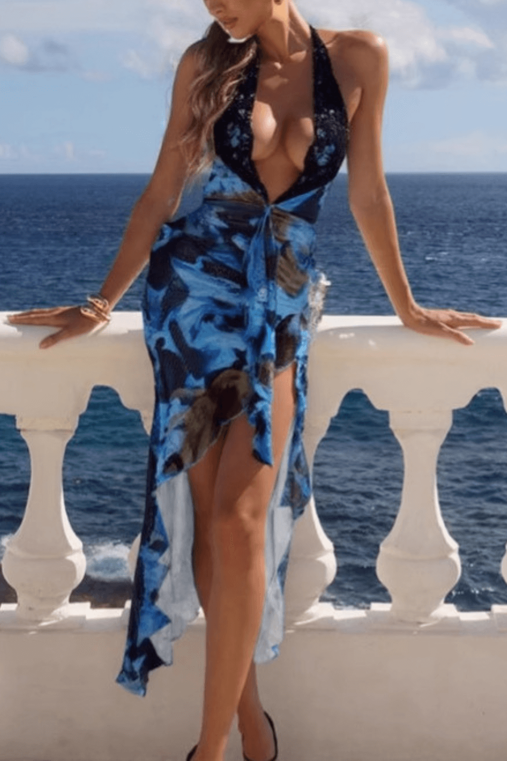 woman in a blue and black floral print halter maxi dress with a plunging neckline and high-low hem standing on a balcony