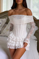 Woman wearing a white off-the-shoulder lace dress with a corset bodice and ruffled skirt, standing by a bed.