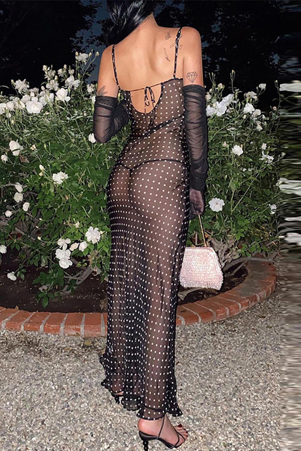 Back view of a woman wearing a black polka dot sheer maxi dress with spaghetti straps and a cowl neck, with black sheer gloves.