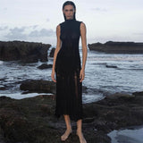 Woman wearing a black sleeveless ribbed knit midi dress with sheer panel detailing standing on rocks by the ocean