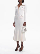 Woman wearing a cream-colored long-sleeved knit dress with a collared neckline, button-up front, dual chest pockets, and a flared hem, holding a white handbag and wearing black heels.