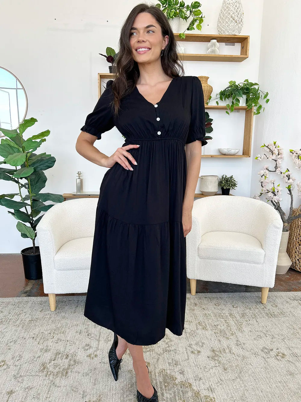 Woman in a black midi dress with short sleeves, smiling and posing with her hand on her hip.