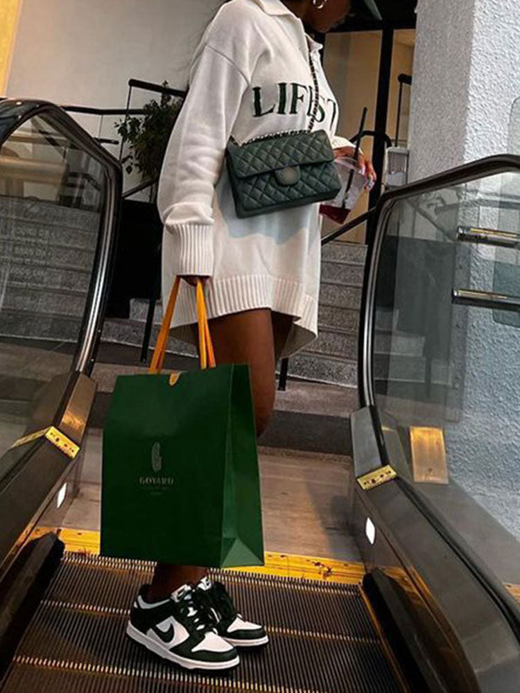 Woman wearing a white oversized knit sweater dress with green 'LIFESTYLE' text graphic, a green quilted handbag, and white and green sneakers, holding a green shopping bag on an escalator