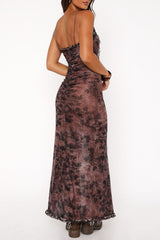Rear view of a woman wearing a brown tie-dye print sheer mesh maxi dress with a high slit and brown boots, posing against a white background.
