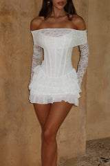 Woman wearing a white off-the-shoulder lace dress with a corset bodice and ruffled skirt, standing against a textured wall.