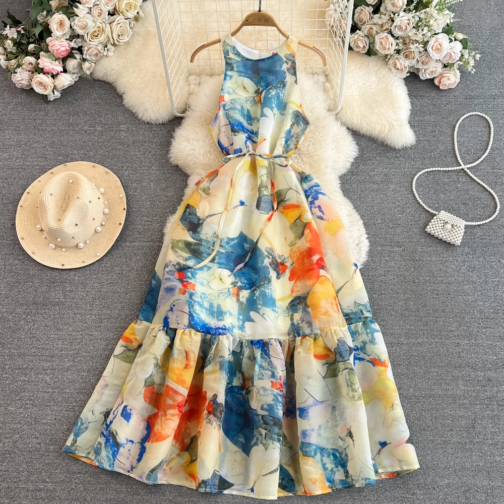 Overhead view of a colorful abstract floral print maxi dress with a tiered skirt and tie waist, laid out with a straw hat and a pearl beaded handbag.