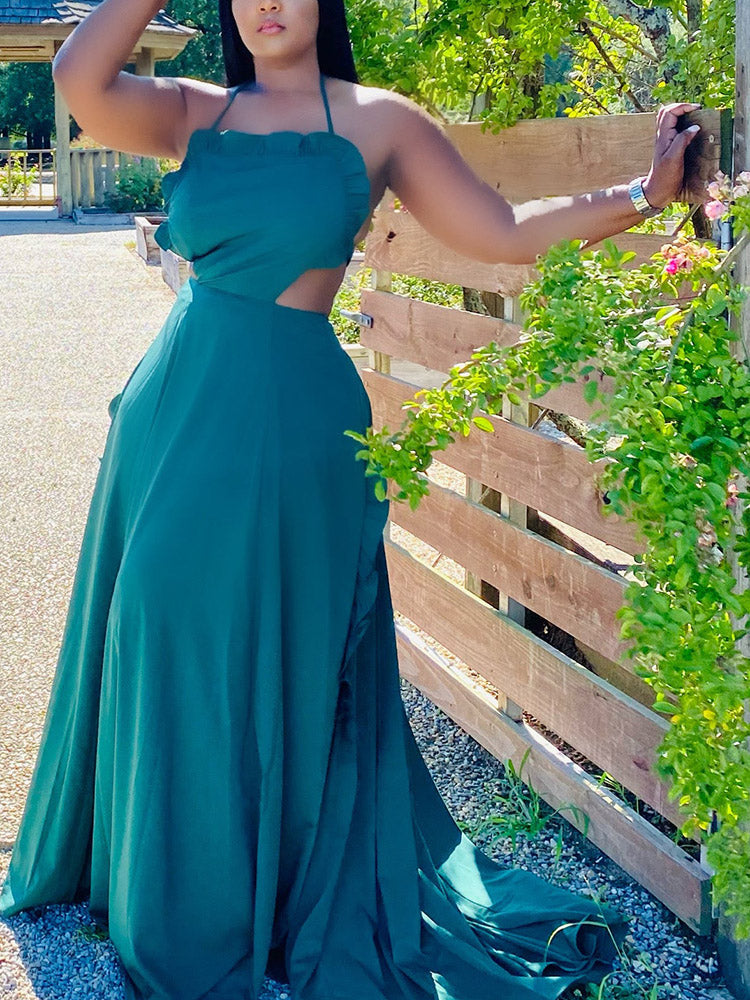 Woman in a teal maxi dress with a halter neckline and cut-out detail, leaning against a wooden fence.