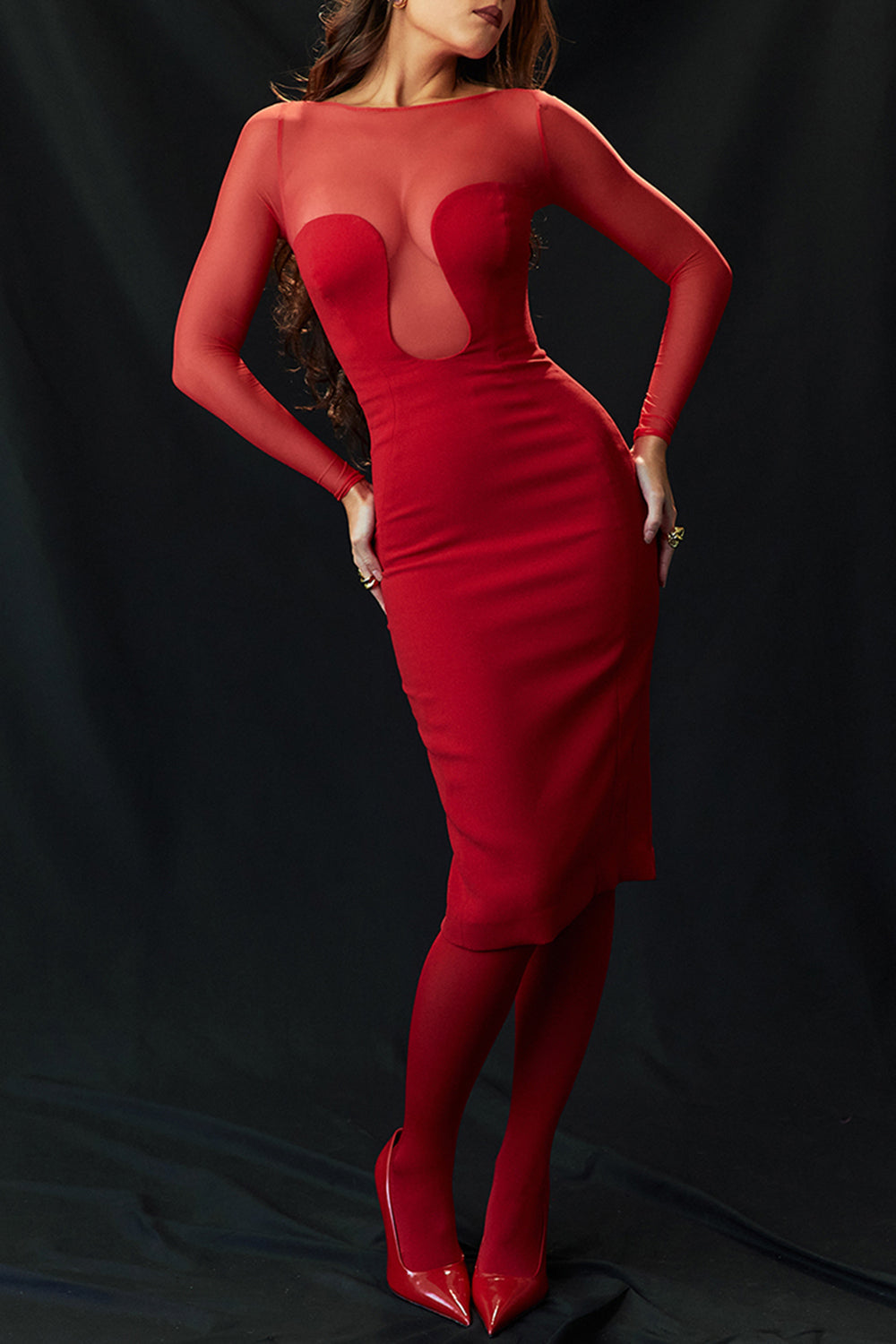 Woman posing in a red sheath dress with sheer illusion neckline and long sleeves against a dark background