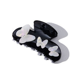 Side view of a black claw hair clip adorned with white iridescent butterfly details and gem accents, casting a shadow.