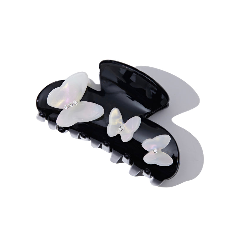 Side view of a black claw hair clip adorned with white iridescent butterfly details and gem accents, casting a shadow.