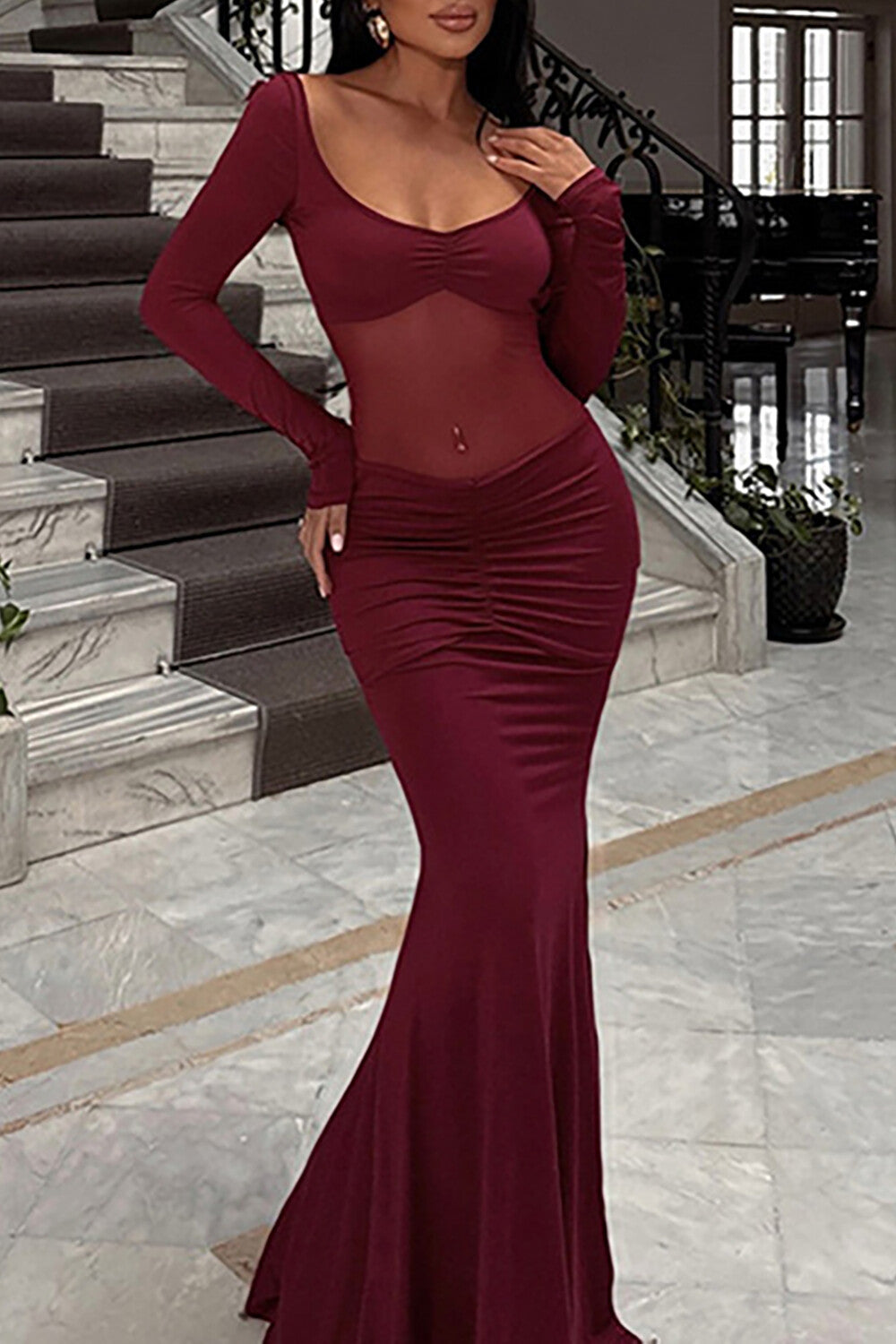Woman wearing a burgundy long-sleeved mermaid dress with a sheer bodice and ruched waist, standing on a staircase.