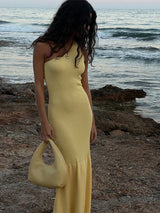 Woman wearing a yellow one-shoulder ribbed knit maxi dress with a mermaid hem, holding a light yellow handbag, standing on a rocky beach near the ocean.