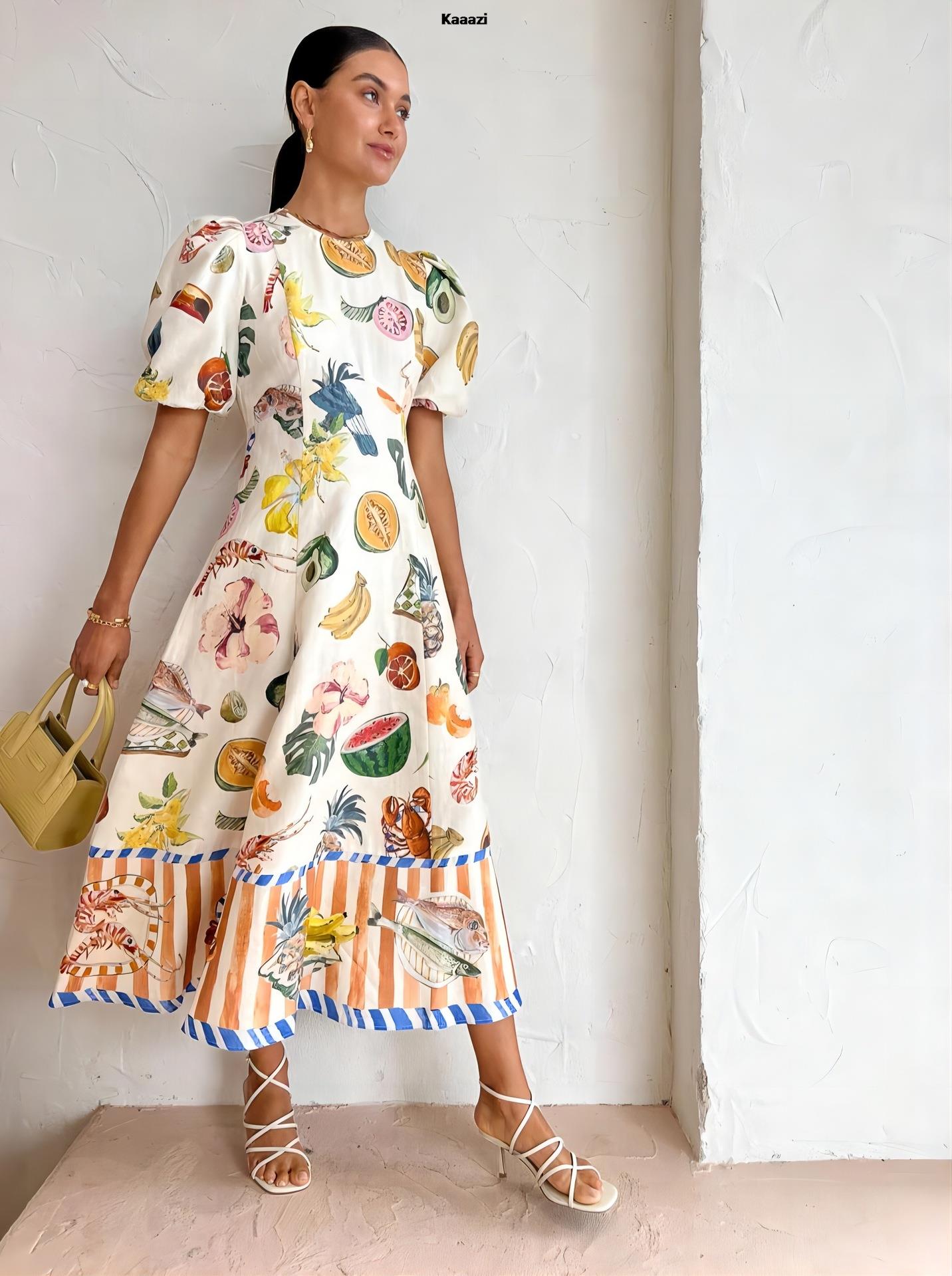 Woman wearing a white midi dress with a colorful tropical fruit and bird print, puff sleeves, and a striped hem with seafood motifs, holding a yellow handbag and standing against a white textured wall.