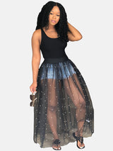 Woman holding sunglasses, wearing a black tank top and a black sheer maxi skirt with white pearl embellishments over denim shorts.