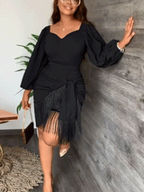 Woman in a black dress with puff sleeves and fringe detail, leaning against a wall.