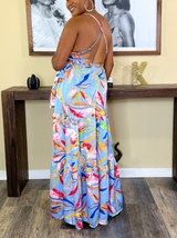 Rear view of a woman wearing a light blue maxi dress with a tropical floral print, showing the criss-cross back straps and tiered skirt design.