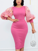 Woman wearing a pink midi sheath dress with sheer puff sleeves adorned with floral appliqués, holding a silver clutch.