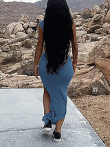 Rear view of a woman wearing a blue one-shoulder, draped maxi dress with a high slit and sneakers, walking away.