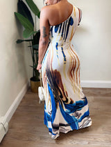 Back view of a woman wearing a one-shoulder maxi dress with an abstract print, holding a white handbag.