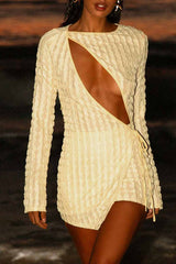 Woman wearing a light yellow textured mini dress with a plunging neckline and long sleeves standing on a beach at dusk.
