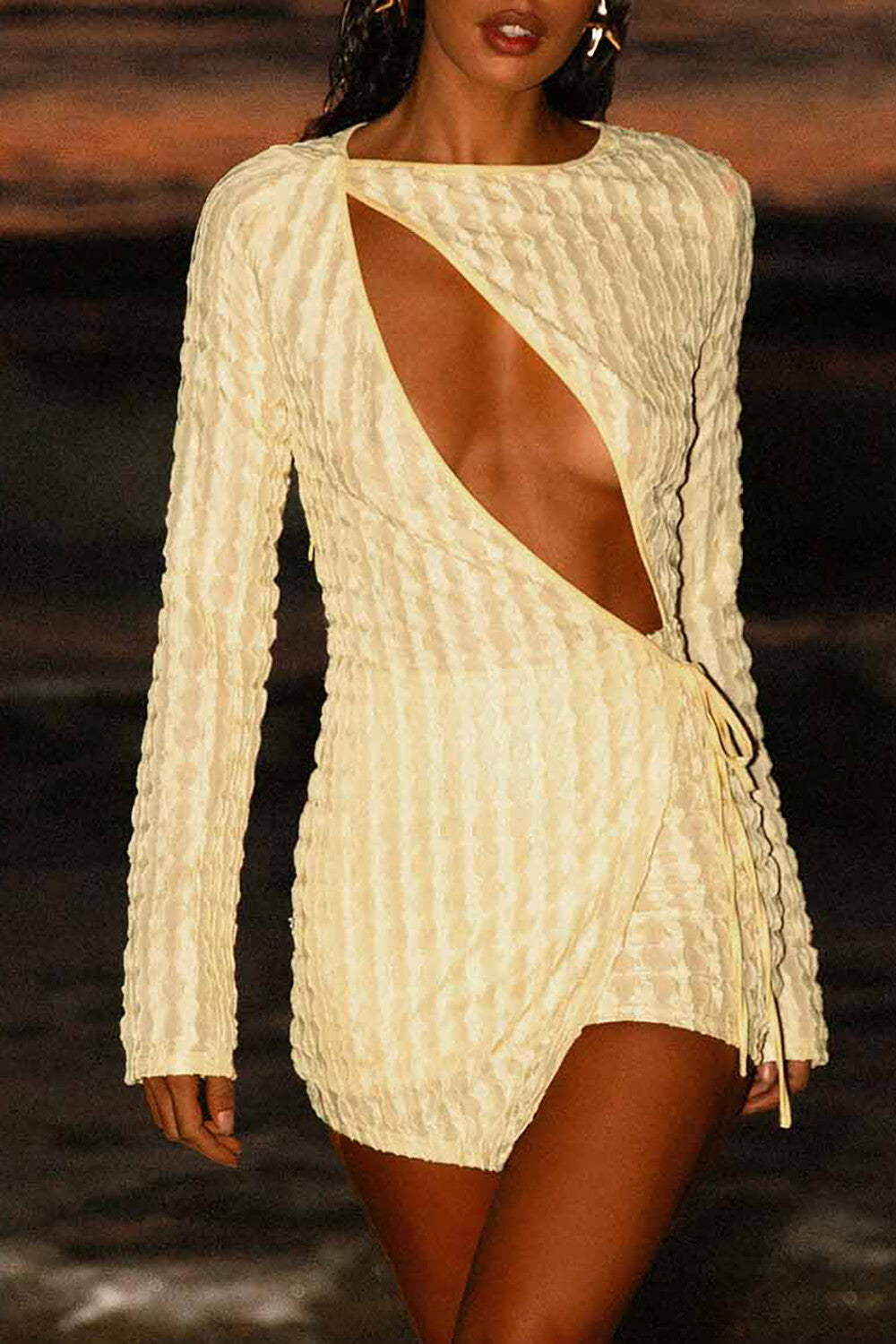 Woman wearing a light yellow textured mini dress with a plunging neckline and long sleeves standing on a beach at dusk.