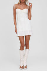 Woman wearing a white textured crinkle mini dress with tie straps and ruffled hem, standing against a white background and wearing white boots.