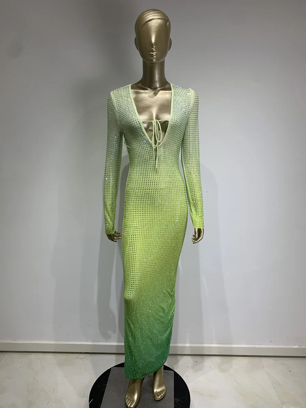 A mannequin displays a long, gradient green dress with long sleeves and a plunging neckline, embellished with rhinestones.