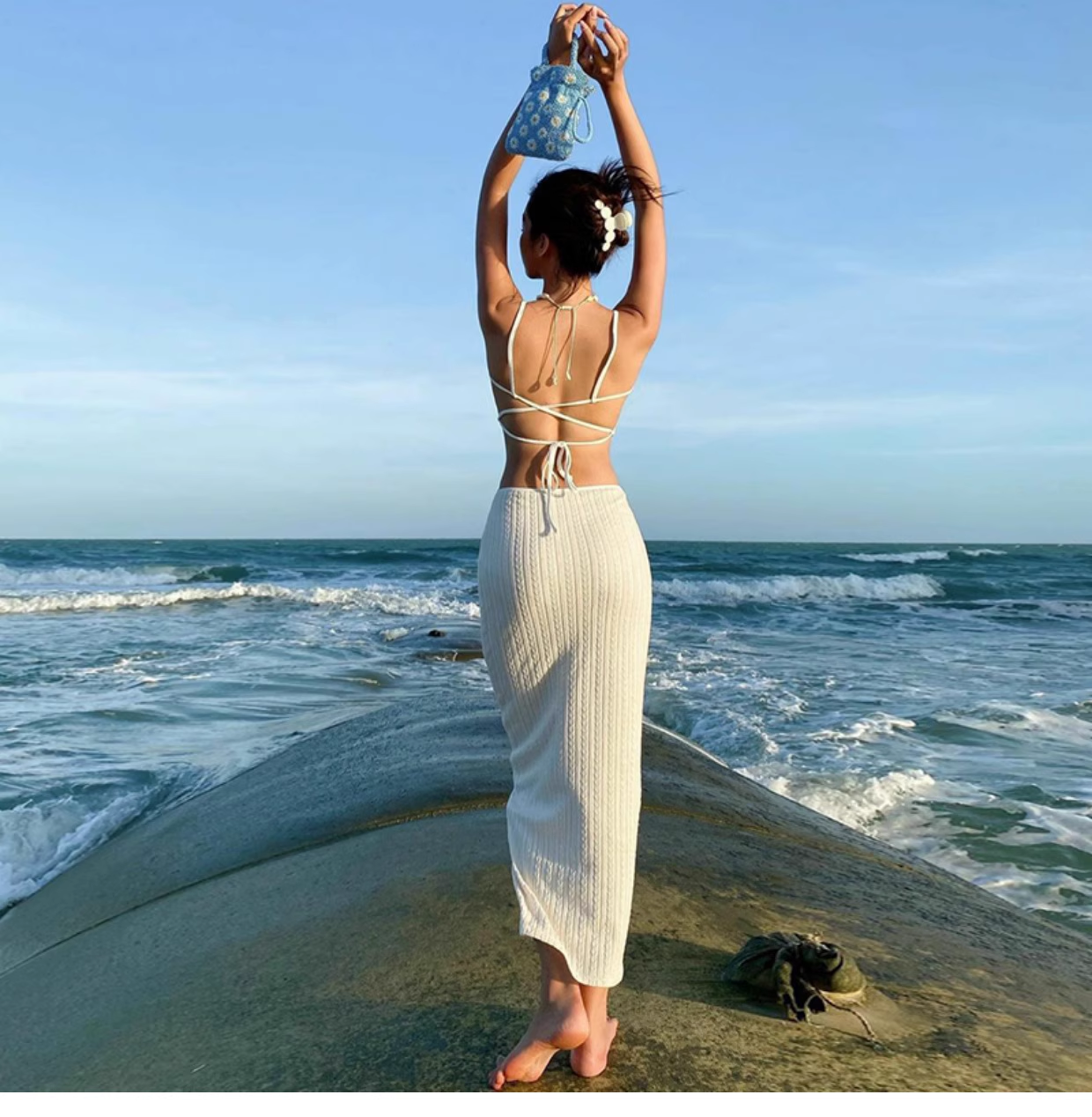 Rear view of a woman wearing a white knit midi dress with cut-out details and a small blue bag, standing by the ocean.