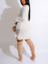 rear view of a woman in a white distressed knit mini dress with an off-the-shoulder neckline and long sleeves, with beige knee-high boots