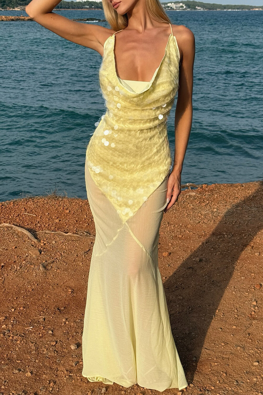 A woman stands on a rocky shore wearing a flowing yellow maxi dress with sequin embellishments.