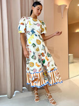 Woman wearing a white midi dress with a colorful tropical fruit and bird print, puff sleeves, and a striped hem with seafood motifs, standing indoors with a curtain in the background.