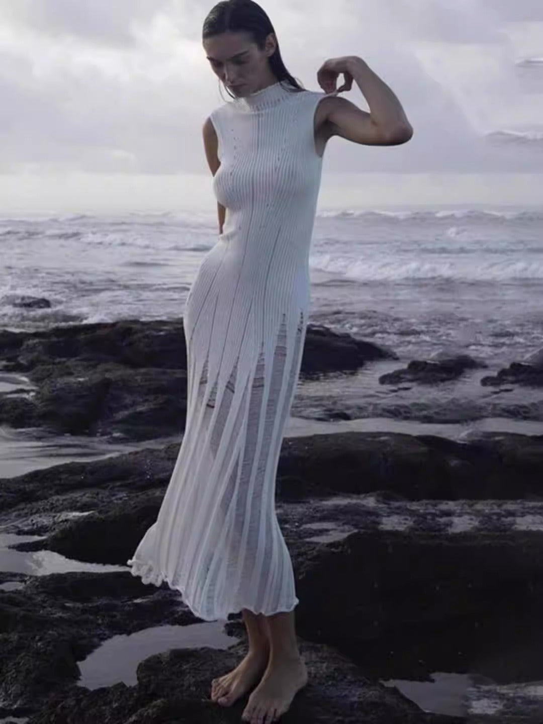 Woman wearing a white sleeveless ribbed knit midi dress with sheer panel detailing posing on rocks by the ocean