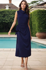 Woman in a navy blue sleeveless cowl neck midi dress with a self-tie belt, walking by a pool.
