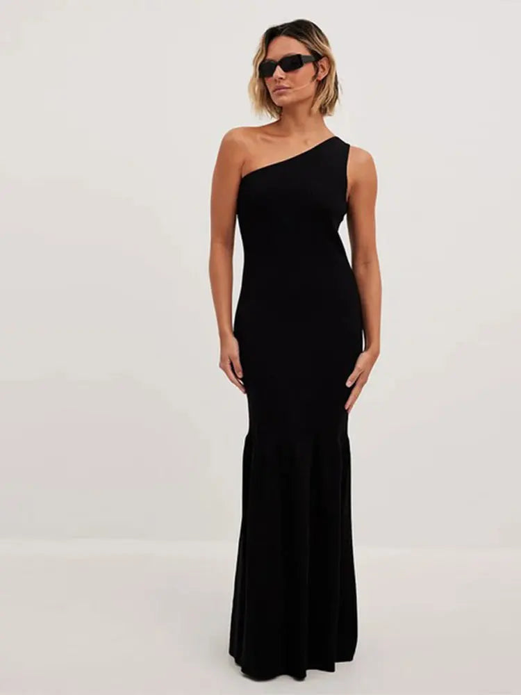 Model wearing a black one-shoulder ribbed knit maxi dress with a mermaid hem, standing against a white background, wearing sunglasses.