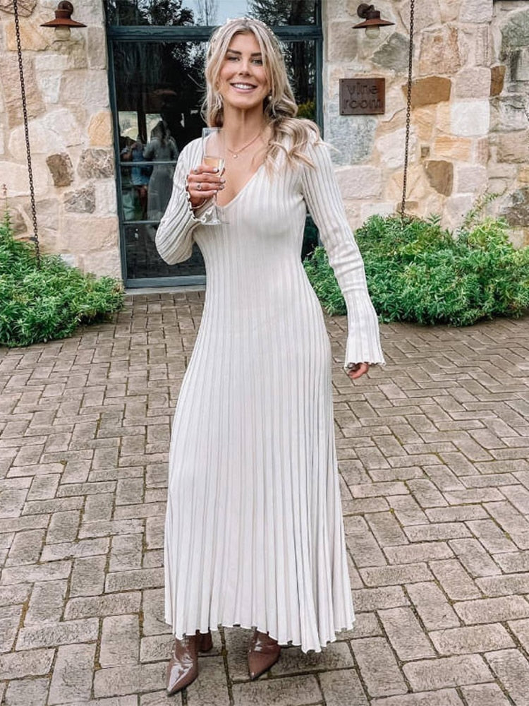 Woman wearing a cream ribbed knit maxi dress with flared cuffs and brown boots, holding a glass of champagne.