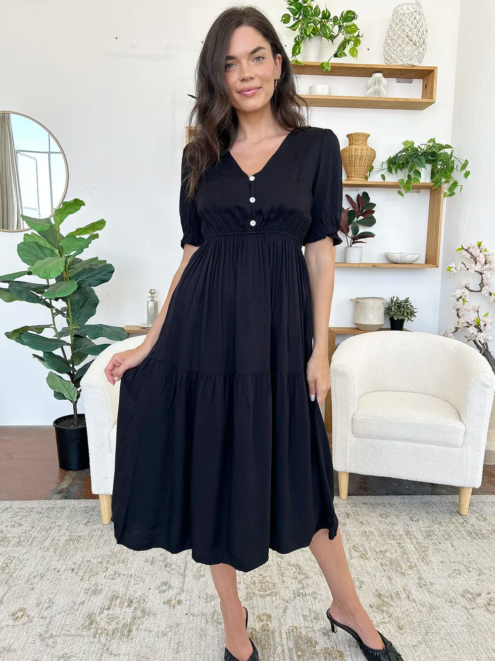 Woman wearing a black midi dress with short sleeves and a tiered skirt, standing in a bright room with plants.