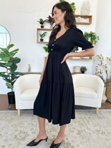 Side view of a woman in a black midi dress with short sleeves, tiered skirt, and V-neckline, posing with her hand on her hip.