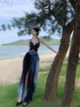 Woman wearing a long, flowing ombre dress with a halter neckline and cutout waist, standing on a grassy area near a beach.